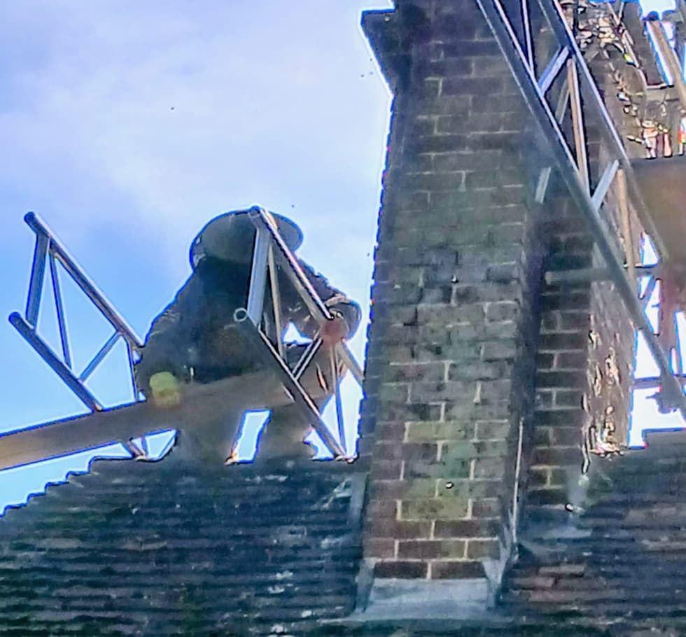 A brick chimney stack reached from the scaffold, working level with the pot above a moss-covered tiled roof