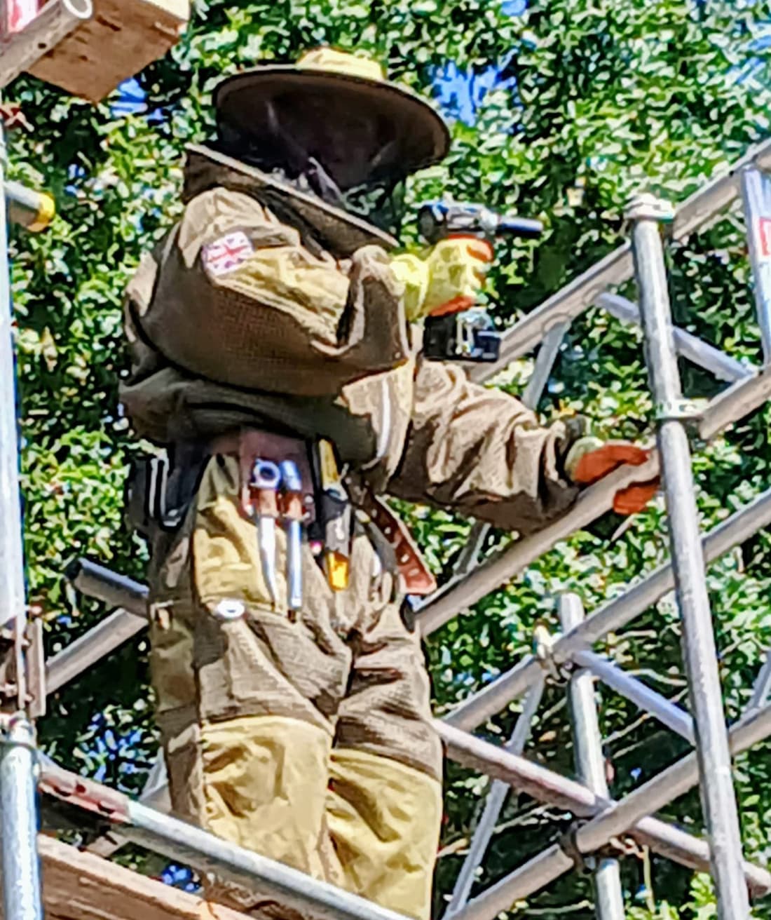 Close view of the pest operative in a full bee suit, veil and tool belt, working from the boarded lift of the scaffold