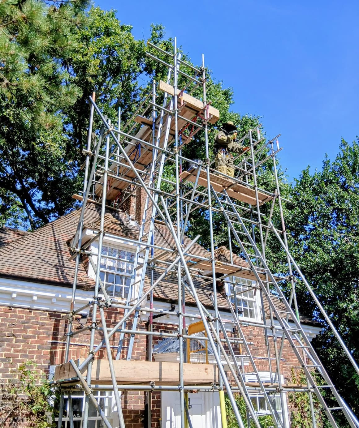 A pest operative in a full bee suit working at a chimney stack from the top lift of an I.C. Scaffolding tower, built up the side of a large detached house